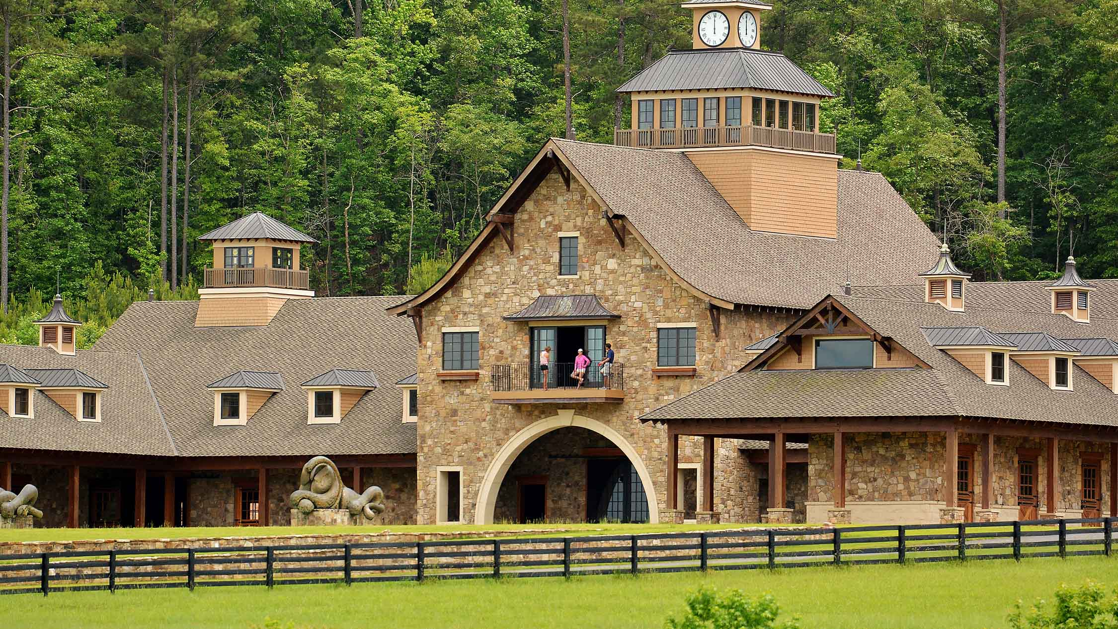 The stone-and-timber equestrian building and clock tower at Bouckaert Farm, framed by woodland and a fenced lawn.